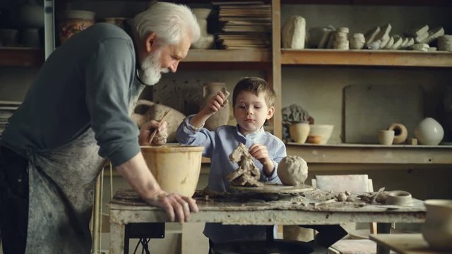 Cute child is playing with clay and potter's tools making ceramic toy in his grandfather's workshop. Grey-haired bearded man is looking at him with love and care and talking.