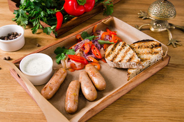 Top view, close up. Grilled sausages with vegetables, tartar sauce on wooden box on gray background. Pepper, tomato, onion, garlic and greens are served nicely on a plate. Restaurant kitchen