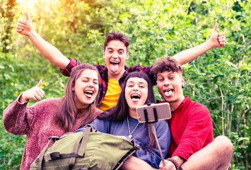 Group of young friends taking selfie with stick on  green forest background - Teenagers having fun using mobile phone technology for self photo on camping excursion day 