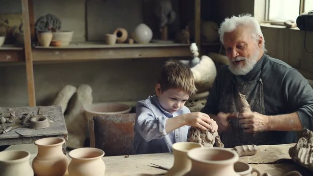 Cute boy is having fun playing with clay in his grandfather's pottery studio while elderly man is kneading clay preparing to mold utensils. Family spending time together concept.