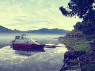 two motor boats moored to the shore of a lake with calm water in an early foggy morning