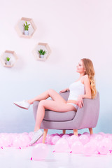 Obraz na płótnie Canvas Sporty girl in white underwear and white sneakers sitting on the purple armchair, smiling and looking at the plants on the wooden shelves in her room with pink balloons on the floor