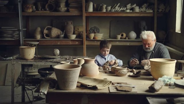 Pottery master is making clay balls preparing to form ceramic figures and showing them to his grandchild who is playing with clay. Handmade clayware and cozy workshop are visible.