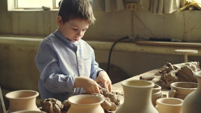 Curious little boy is playing with clay at working table in potter's workshop, kneading clay and tearing into pieces. Childhood, hobby, new experience concept.