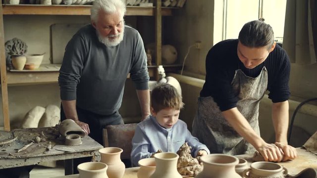 Professional potter is kneading clay on worktable in home studio while his son is helping him and his elderly father watching them from behind. Small family business concept.