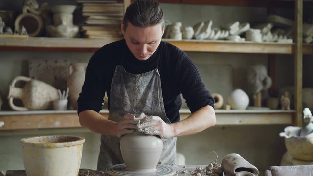 Experienced ceramist is forming ceramic vase from clay on spinning throwing wheel while working in potter's workshop. Professional is focused on work.