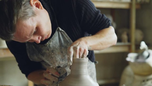Close-up shot of young potter molding ceramic vase from clay on throwing wheel while working in potter's workshop. Shelves with handmade eathenware in background.