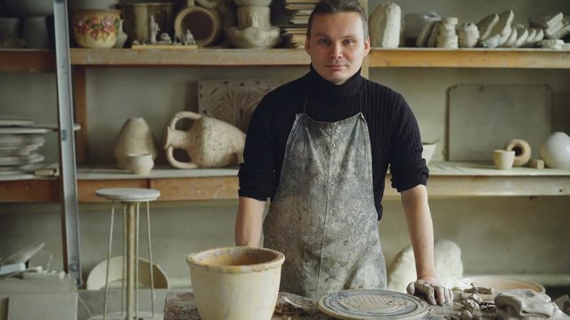 Portrait of handsome young potter in muddy apron standing at table in workshop and looking at camera. Shelves with handmade vases and pots in background.