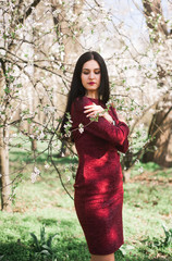 beautiful young brunette with long hair in a park on a spring in the middle of flowering trees in a red dress