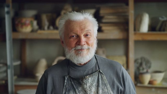 Close-up portrait of senior potter in muddy apron standing at table in workshop and looking at camera. Shelves with handmade vases and pots in background.