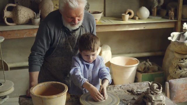 Young cute boy is making clay figure on throwing wheel while his grandfather experienced sculptor is standing near worktable and watching carefully. Family tradition concept.