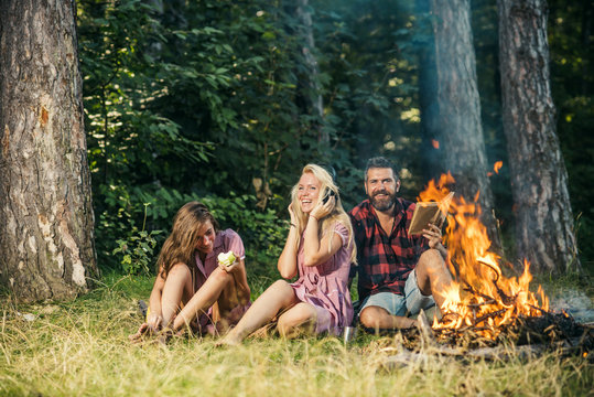 Happy friends having fun at picnic in woods. Bearded man with his girlfriend and her sister sitting near campfire. Blond girl listening to music while her friend is eating apple