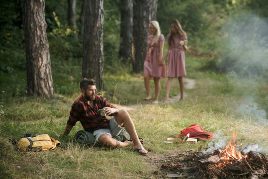 Picnic at sunny meadow. Two girls wandering in woods while thoughtful man is sitting on grass. Brutal bearded man drinking coffee or tea by campfire. Friendship and nature concepts