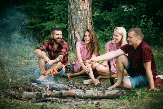 Young couples having picnic in woods. Bearded man and his best friend frying sausages over fire. Happy people sitting around campfire, friendship and leisure concept