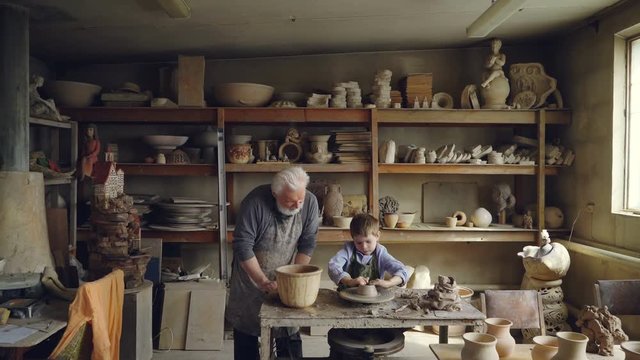 Skilled child is forming pot on potter's wheel while working with his grandfather in his figuline home studio. Professional equipment and beautiful ceramic things in background.