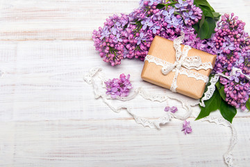 Obraz na płótnie Canvas Prezent z kokardą otoczony kwiatami bzu na białym drewnianym tle, miejsce na tekst. Świąteczna kompozycja z okazji Dnia Matki, urodzin lub rocznicy. 