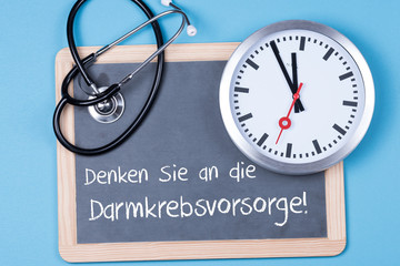 Denken sie an die Darmkrebsvorsorge als Text auf einer Tafel und einer Uhr mit Stethoskop
