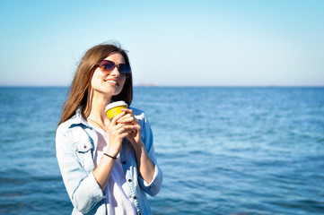 Beautiful young woman in sunglasses and with a cup of coffee smiling
standing by the sea