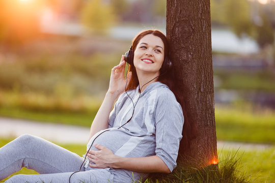 Young pregnant woman listening to music from a smartphone on a lawn outdoors