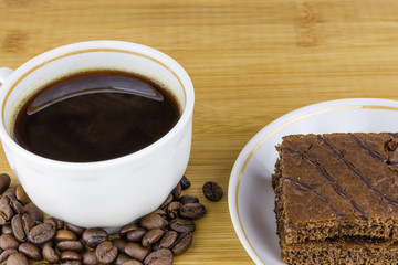 Cup with coffee, coffee beans, cake on a saucer against the background of a bamboo wooden board, morning breakfast close-up