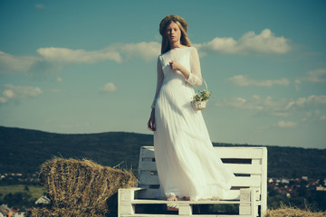 Fototapeta na wymiar Albino girl with flowers, natural beauty. Woman bride in wedding dress on wooden bench. Sensual woman in wreath on long blond hair. Fashion model at sunny blue sky on vacation