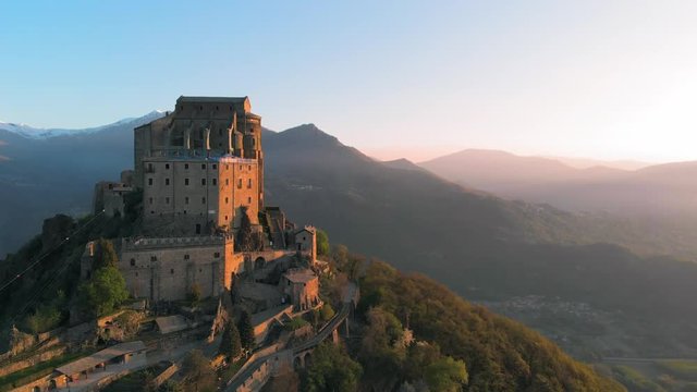 Aerial: drone flying at old medieval abbey perched on mountain top, background snowy Alps at sunrise. Sacra di San Michele (italian) - Saint Michel Abbey (english traslation) - Turin, Italy 