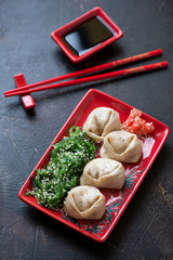 Red tray with steamed asian dumplings, seaweed salad and dipping sauce. Studio shot on a dark brown...