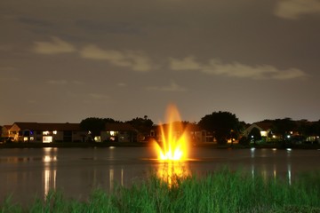 Yellow Golden Glowing Illuminated Fountain at Night on a Lake Overlooking a Residential Community with House Lights On