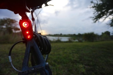 Fototapeta na wymiar Closeup of Red Bicycle Tail Light Illuminated on Bike with Green Grass, Trees and Lake Out of Focus in the Background Beneath a Cloudy Overcast Sunset