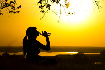 silhouette of a woman drinking water after fitness training in nature, female profile at sunset, concept of sport and relaxation