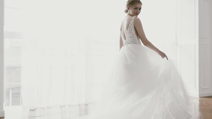 Beautiful bride in a gorgeous dress and bridal veil stands by the window dreaming, smiling charmingly. Happy moments, couple goals, morning of a bride