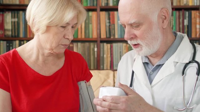 Male professional doctor in white coat with stethoscope at work. Senior man physician measuring pressure to sick senior female patient by tonometer at home. Consulting about treatment and therapy