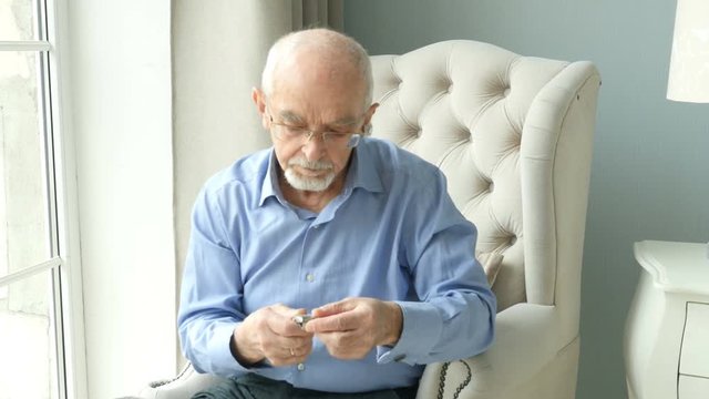 Elderly man sits on a chair by the window and puts on a watch