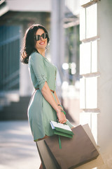 woman in summer tunic and eyeglasses is standing and smiling with shopping bags in her hands in the city
