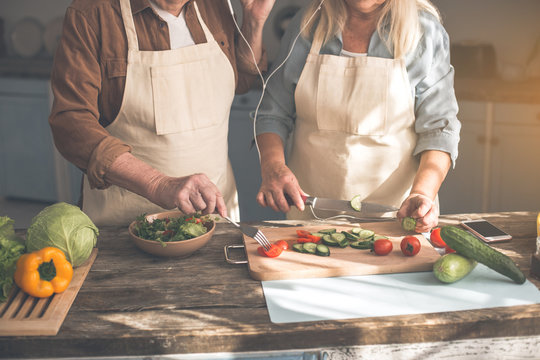 Close up of male and female arms making salad in kitchen. Mature man and woman are standing at desk while wearing earphones