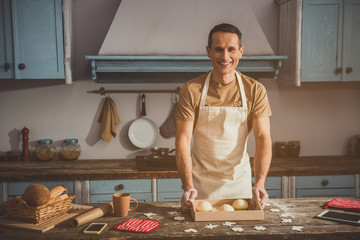 Portrait of glad guy standing at the kitchen table and looking at camera. Tray with jelly is on desk. Copy space in right side