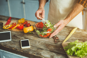 Close up of man arms intermixing vegetable ingredients in glass bowl. Tablet and smartphone are on desk