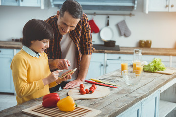 Father and son standing at the kitchen table. They are looking at mobile phone with joy. Vegetables...