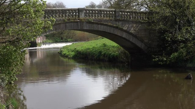 Water flowing over a distant weir viewed through an ornate stone arched bridge with decorative parapet.