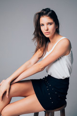 Young brunette woman fashion model sitting on chair isolated on gray background