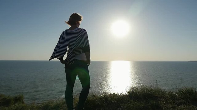Back view of a beautiful blond woman in a striped sea pareo keeping hands on her hips and looking at a golden path at sunset in summer in slo-mo