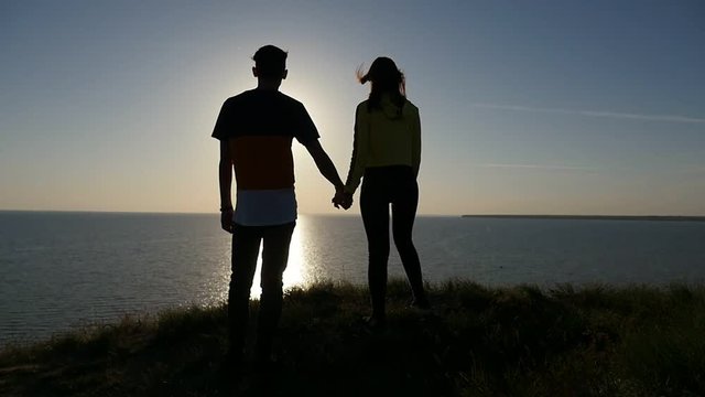 Back view of a stylish man keeping the hand of his happy girlfriend at a gorgeous sunset with a shining sun path at the Black Sea in slo-mo