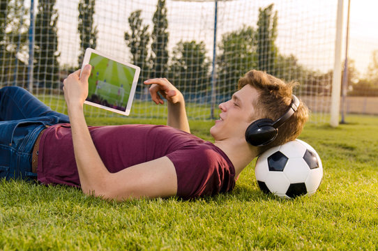 Teenager laying on grass with soccer ball behind his neck , watching soccer game on tablet 