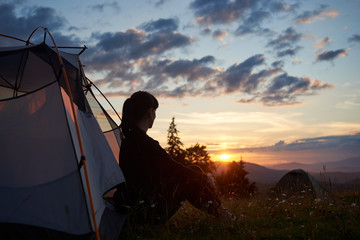 Woman tourist sitting in tent at the top of mountain enjoying the sunset on blurred background of mountains and hills under incredible beautiful evening sky with clouds. Concept of healthy lifestyle.