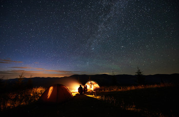 Rear view of silhouette mother and two sons hikers at camping in mountains, sitting on log beside campfire, two tents, looking at amazing night sky full of stars and Milky way, enjoying evening scene
