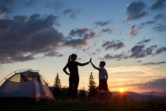 Silhouette of a young woman and a child giving each other a high five near a camping at dawn on top of a mountain. A beautiful landscape of fir-trees and sun rising over the mountains and hills