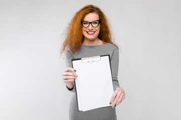 Naklejka premium Charming young girl with red hair. A young girl in a gray sweater. Young girl on a gray background. A young girl with glasses holds a pen and a notebook in her hands