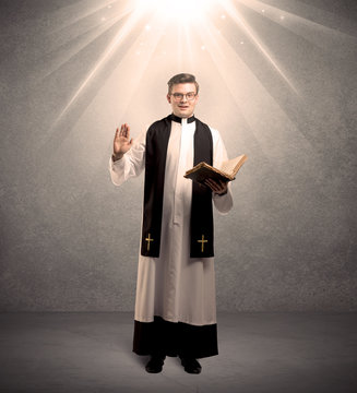 A male religious young priest in black and white dress giving his blessing, holding the holy bible while being illuminated from strong light beams coming from above concept