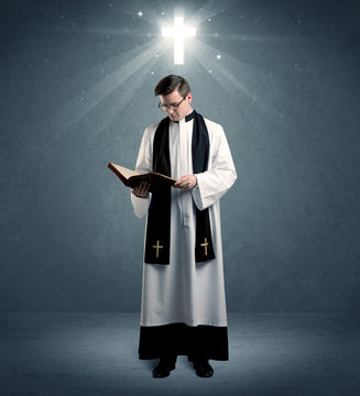 A young caucasian priest with deep faith blessing with the holy bible in his hand in front of blue wall background with illustrated glowing cross concept.