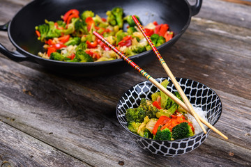 wok with vegetables, bowl of rice and chopsticks on wooden ground